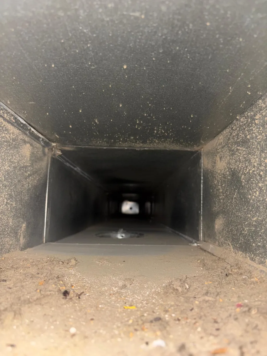 Inside a rectangular supply duct with dust and debris along the bottom, before cleaning